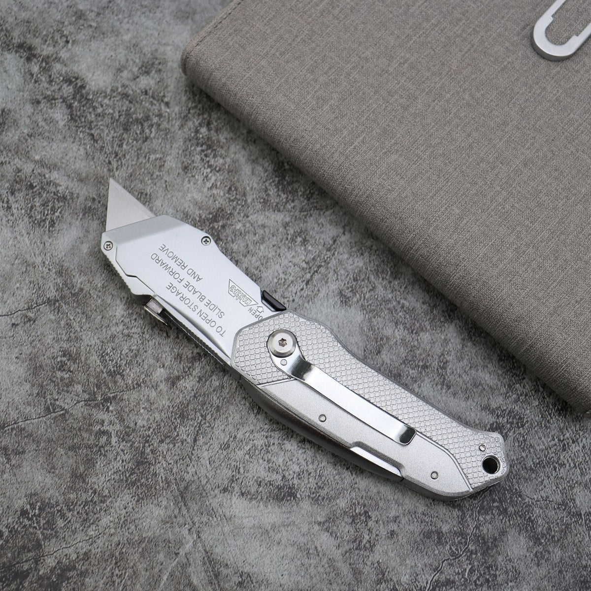 silver folding utility knife with gray storage case on stone surface